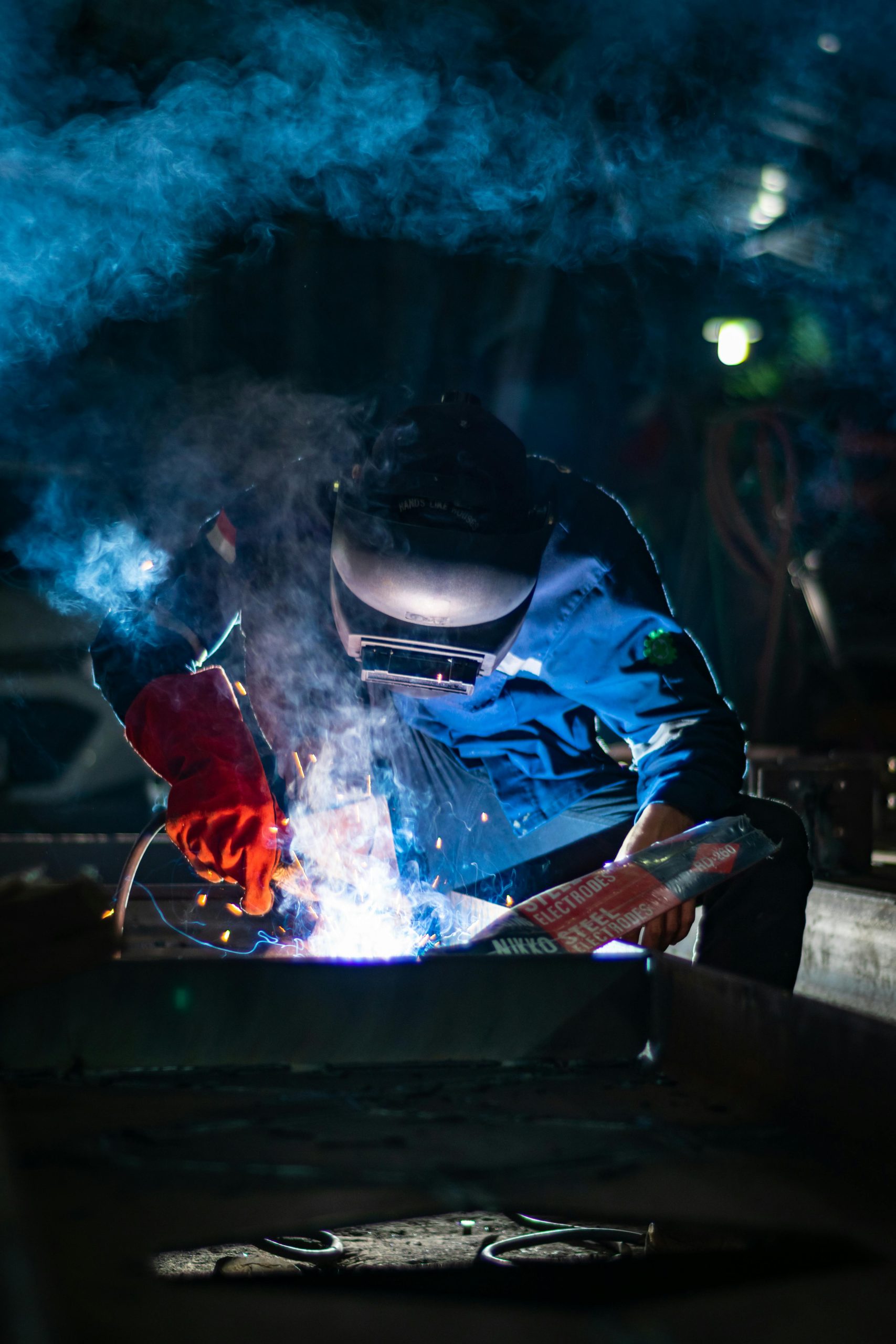 A welder engaged in intense work, surrounded by sparks and smoke, creating a dramatic industrial atmosphere.
