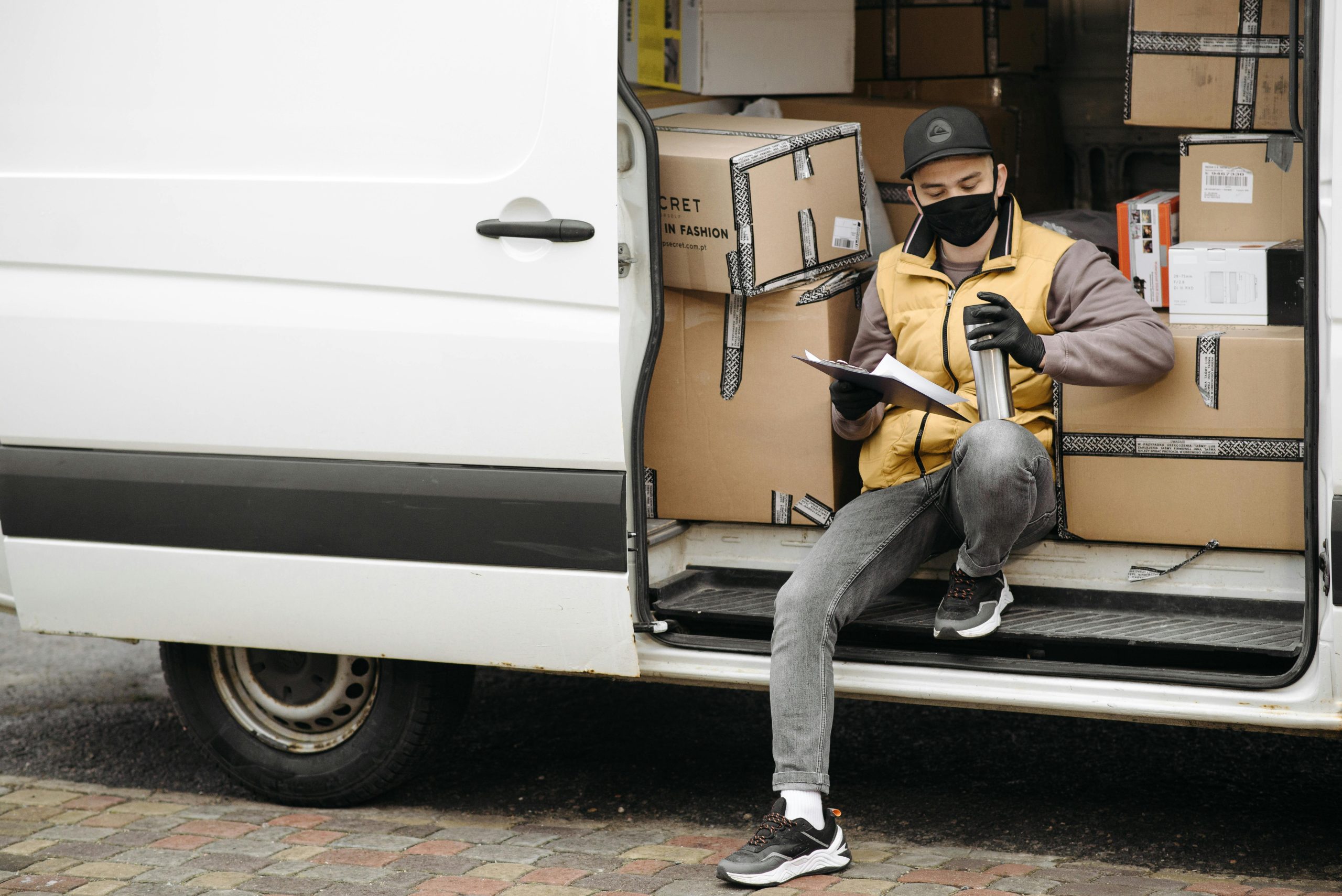 Masked courier sitting in a van, reviewing documents, surrounded by delivery boxes.